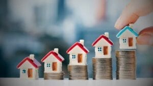 Tax Implications for Foreigners Buying Property in Mexico - Sell Business A hand placing a miniature house atop ascending stacks of coins, symbolizing increasing real estate values in Mexico against a blurred city background. Exit Advisor Business Broker