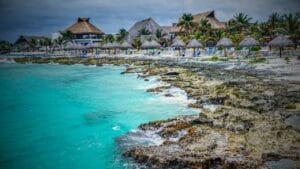 What to Expect From the Mexico Real Estate Closing Process - Sell Business Rocky shoreline with turquoise sea water in front of a beach resort with thatched-roof huts under an overcast sky, ideal for foreign investors. Exit Advisor Business Broker
