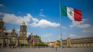 The Importance of Due Diligence in Mexico's Real Estate Transactions - Sell Business A large Mexican flag waves in the breeze over a busy public square flanked by ornate buildings under a clear blue sky attracting foreign investors. Exit Advisor Business Broker