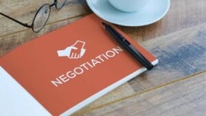 How to Negotiate the Terms of Your SBA Loan- Sell Business A book titled "Loan Negotiation" with a handshake icon on the cover, alongside a pen, glasses, and a coffee cup on a wooden table. Exit Advisor Business Broker