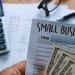 Navigating SBA Loans for Small Business Purchases - Sell Business A person holding a "small business" flyer over a U.S. tax form, with a calculator, glasses, and money nearby on a desk. Exit Advisor Business Broker