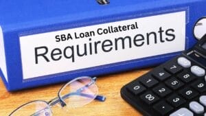 SBA Loan Collateral Requirements: A Detailed Look- Sell Business Blue binder labeled "SBA loan collateral requirements" beside a calculator and glasses on a wooden desk. Exit Advisor Business Broker