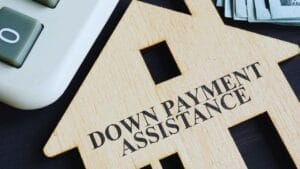 SBA Loan Down Payment Requirements Explained - Sell Business Wooden house-shaped cutout with "SBA loan down payment assistance" text, placed near a calculator and stacks of money on a desk. Exit Advisor Business Broker