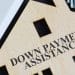 SBA Loan Down Payment Requirements Explained - Sell Business Wooden house-shaped cutout with "SBA loan down payment assistance" text, placed near a calculator and stacks of money on a desk. Exit Advisor Business Broker