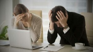 Identifying Causes of Business Failure - Sell Business Two professionals looking distressed at a desk with a laptop and paperwork, each with hands on their heads in a bright office. Exit Advisor Business Broker
