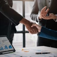 Buying Assets from a Liquidating Company - Sell Business Two business professionals shaking hands at a meeting with others clapping and a digital tablet displaying market forecasting graphs on the table. Exit Advisor Business Broker