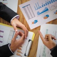 Strategies to Stop Losing Business and Regain Growth - Sell Business Two professionals discussing data charts and reports on a wooden table, pointing at graphs and making notes on forecasting distressed business. Exit Advisor Business Broker