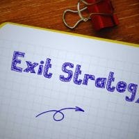 Sell Business Notebook with the words "business exit strategy" written on it, alongside a pen, paper clips, and a yellow notepad on a wooden table. Exit Advisor Business Broker