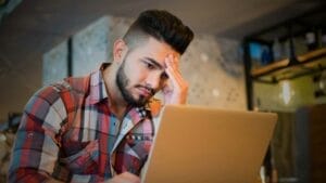 Investing in Distressed Assets: Opportunities & Risks - Sell Business A young man with a beard, wearing a plaid shirt, looks concerned while using a laptop for market forecasting in a warmly lit room. Exit Advisor Business Broker