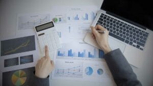 Maximizing Returns from Liquidation Sales - Sell Business An overhead view of a person working at a desk with financial documents, using a calculator and pointing at a laptop screen, surrounded by charts and graphs related to the distressed business market. Exit Advisor Business Broker