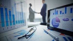 How to Manage a Business Liquidation Process - Sell Business Two business professionals shaking hands behind a blurred foreground of financial charts and graphs, with a focus on glasses and a laptop displaying distressed business market data. Exit Advisor Business Broker