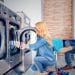 How to Start a Laundromat Business - Expert Tips  - Sell Business A woman loads clothes into a washing machine at the laundromat while a man sitting nearby uses his phone, perhaps reading about the latest business news on franchising costs and profitability. Exit Advisor Business Broker